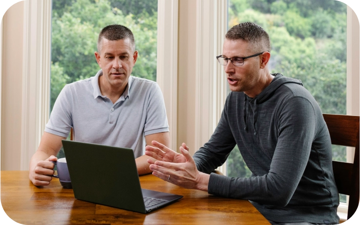 A gay couple sit at their kitchen table, speaking with a doctor on their computer