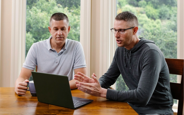 A gay couple sit at their kitchen table, speaking with a doctor on their computer