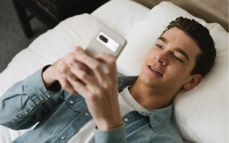 A smiling man reclines on his bed while looking up at his phone.