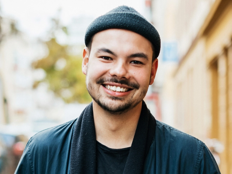A smiling young man with a beard and a dark beanie hat looks directly at the camera. He is wearing a dark jacket and scarf, with a blurred outdoor background.