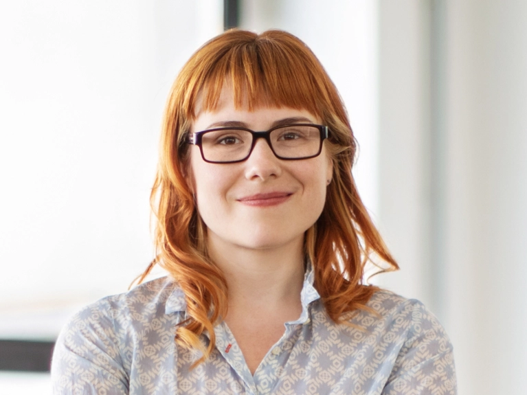 A woman with red hair and bangs, wearing dark-rimmed glasses and a patterned shirt, smiles faintly at the camera. The background is a bright, blurred indoor setting.