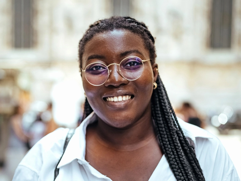 A young Black woman with braided hair and round gold-rimmed glasses smiles broadly at the camera. She is wearing a white collared shirt, with a blurred urban background.