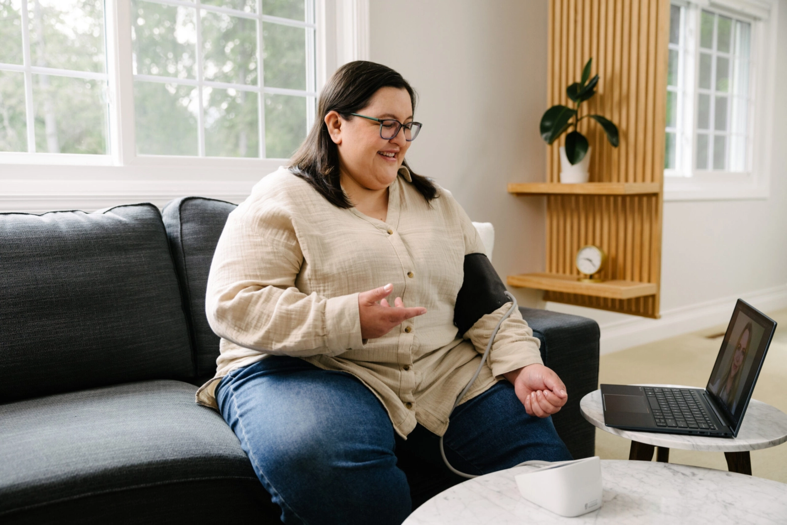 A person wearing glasses and a light-colored shirt sits on a gray couch, using a blood pressure monitor on their arm. They are looking at and gesturing towards a laptop on a small table, which displays a video call with a doctor.