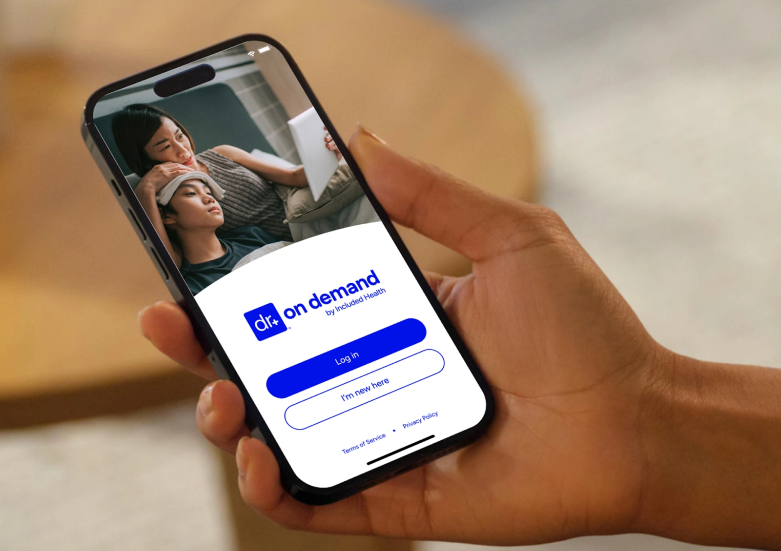 A hand holding a smartphone displaying the "dr on demand by Included Health" app's login screen. The screen also shows a video call between a woman and a child with a compress on their forehead.