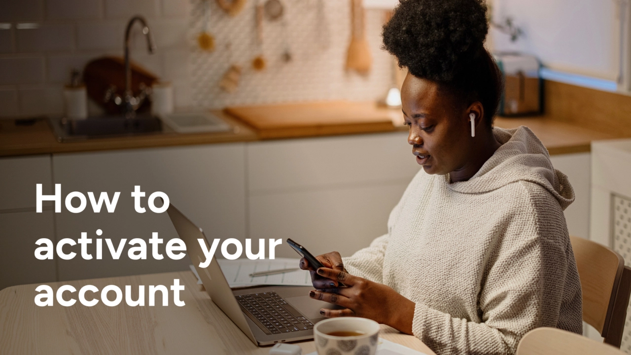 A woman with her hair in a bun, wearing a light gray hoodie and wireless earbuds, sits at a kitchen table. She is looking down at her phone while her laptop is open in front of her. The text "How to activate your account" is overlaid on the bottom left of the image.