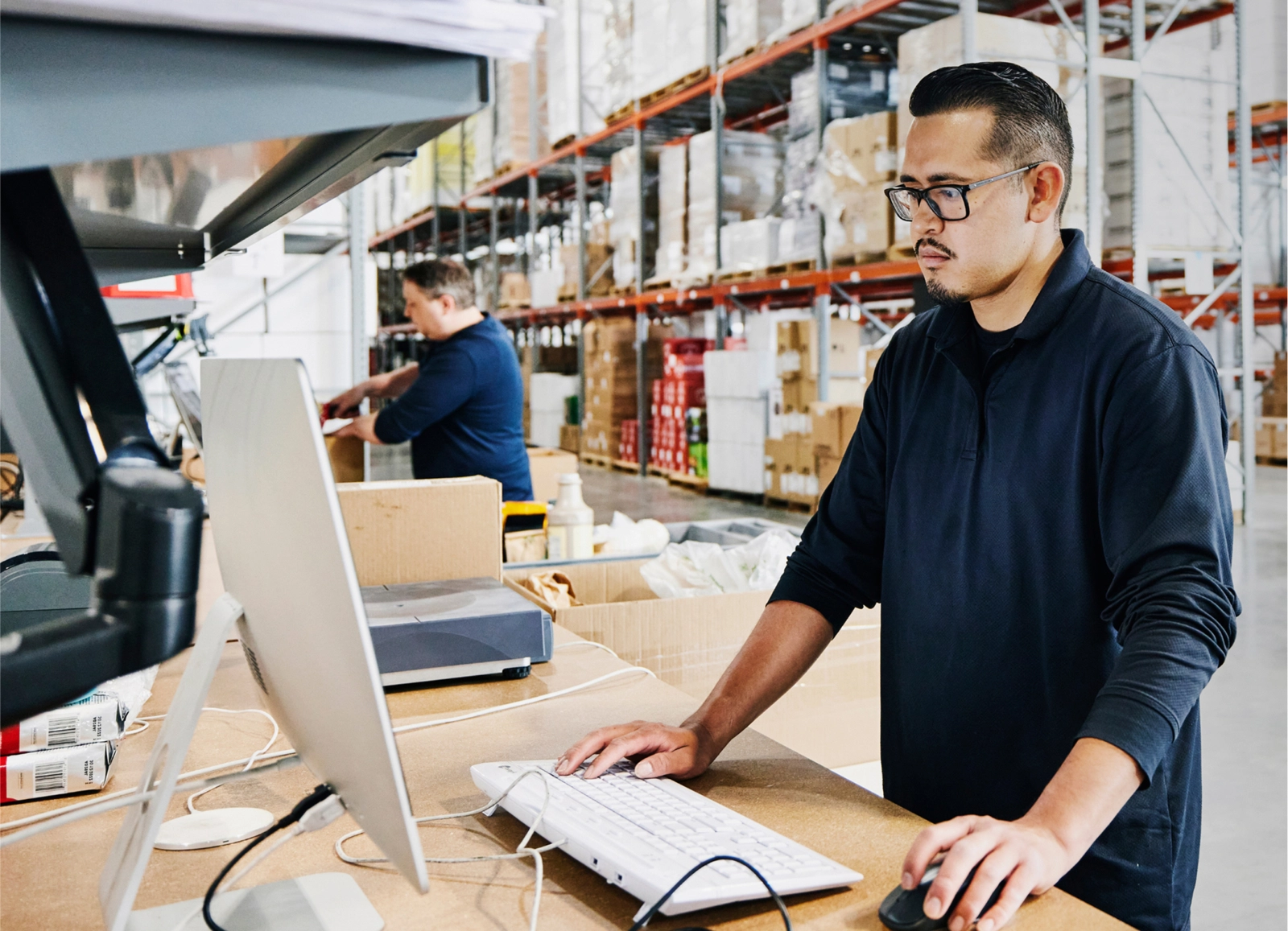 A man with glasses and a mustache, wearing a dark blue polo shirt, works at a computer station in a warehouse. He is using a mouse and keyboard, with shelves of packaged goods and another worker in the blurred background.