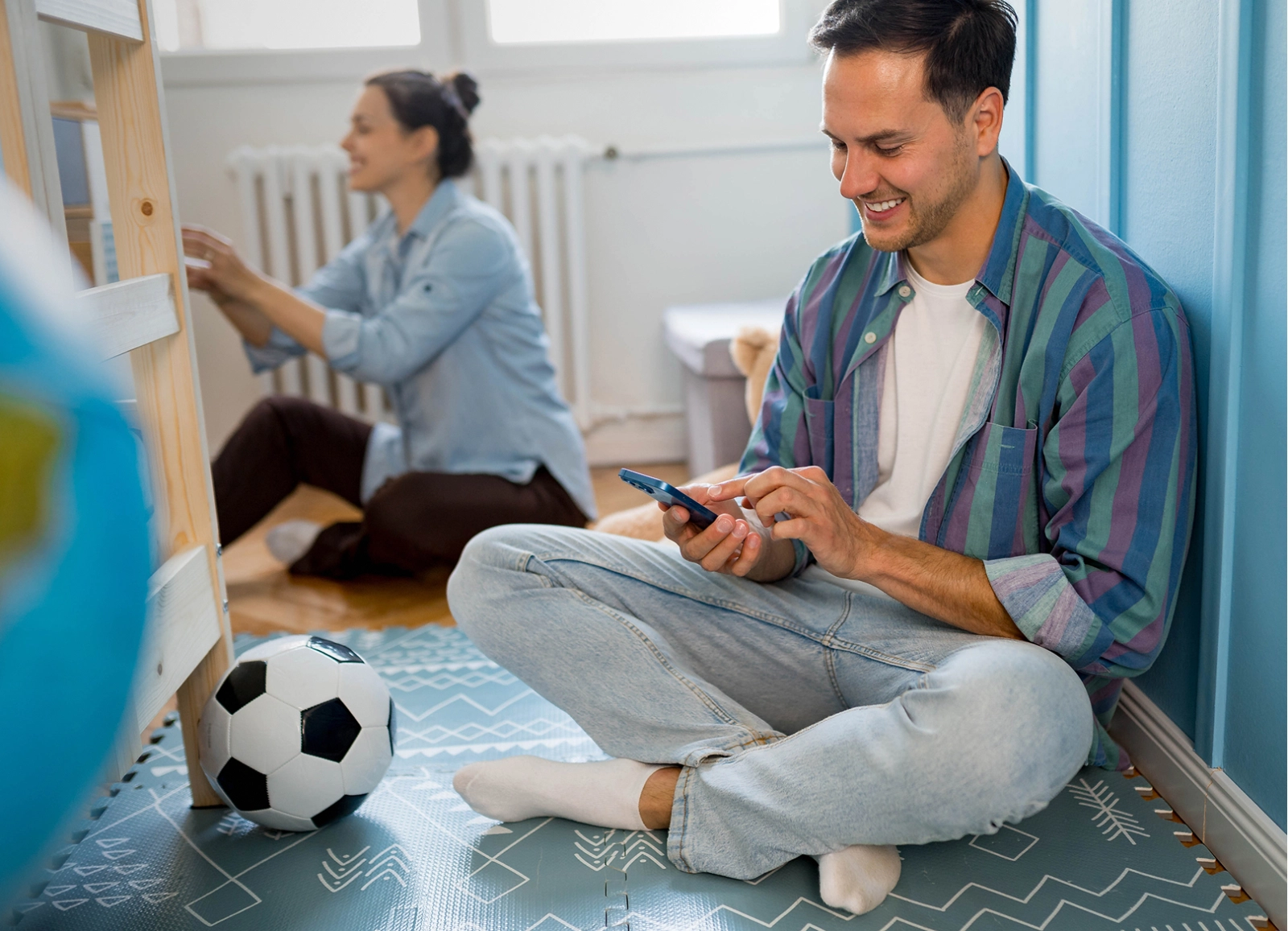 A man sits cross-legged on the floor, smiling at his smartphone, while a pregnant woman works on a piece of furniture in the blurred background. A soccer ball is in the foreground.