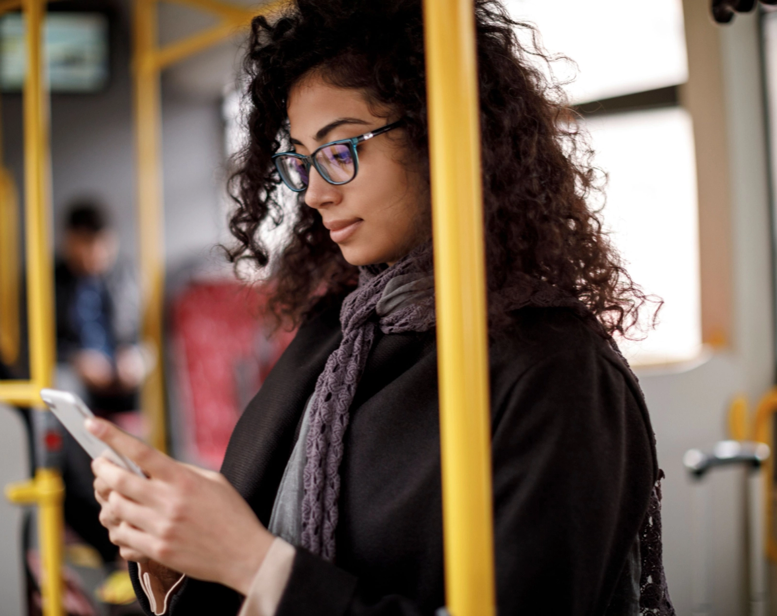 A young woman with dark, curly hair and blue glasses looks intently at her smartphone while riding public transportation. She is wearing a dark coat and a gray scarf.