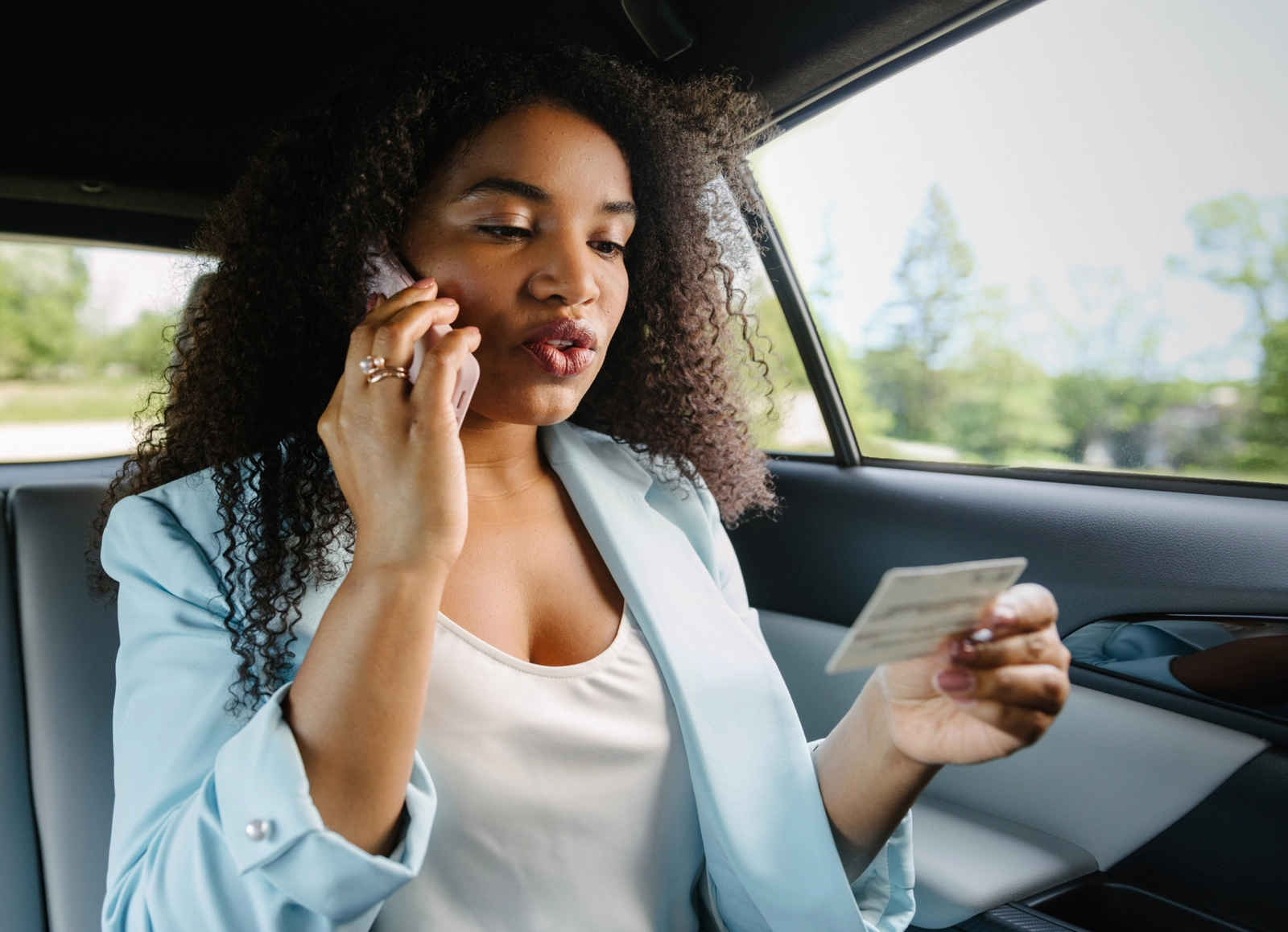 A woman with curly hair sits in the back of a car, holding a cell phone to her ear and looking at her insurance card.
