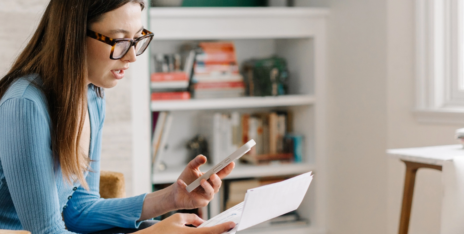 Woman with glasses sits on a couch, holding a smartphone in one hand and a stack of medical bills in the other. She appears to be looking at her phone, possibly using it to resolve a billing issue.