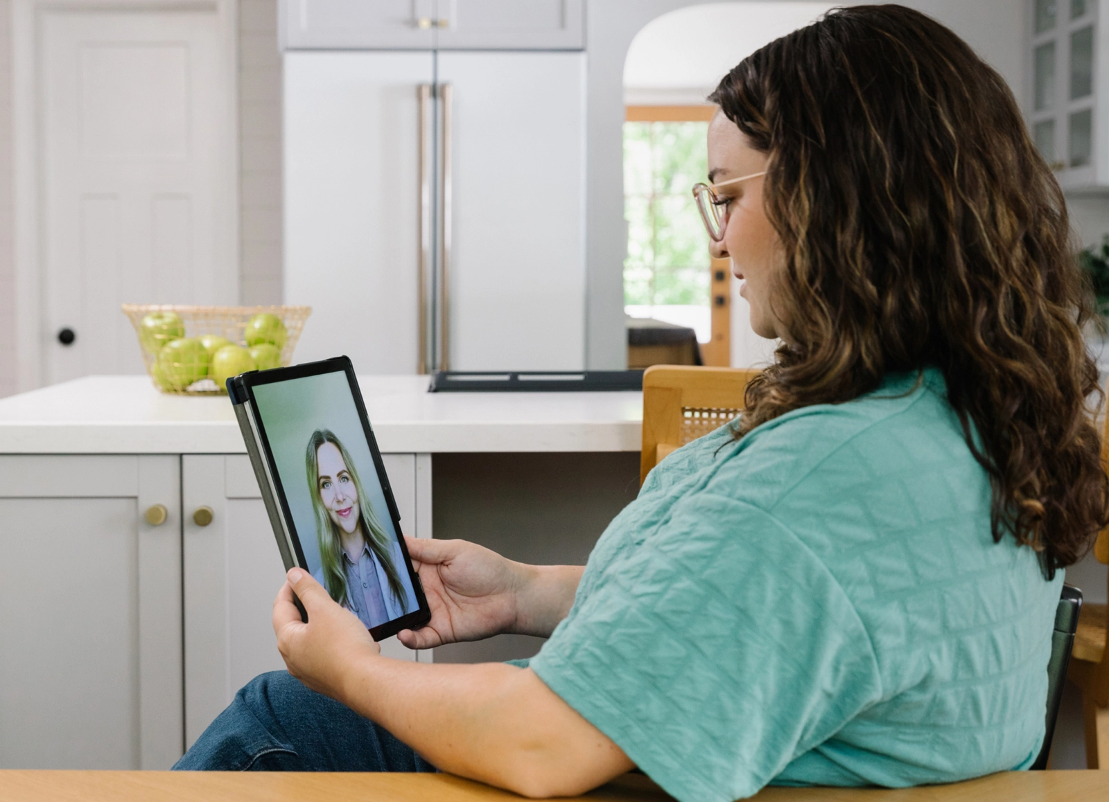Alt Text: A woman with long, curly brown hair and glasses sits at a kitchen table. She is looking at a tablet, which is showing a video call with a medical professional.