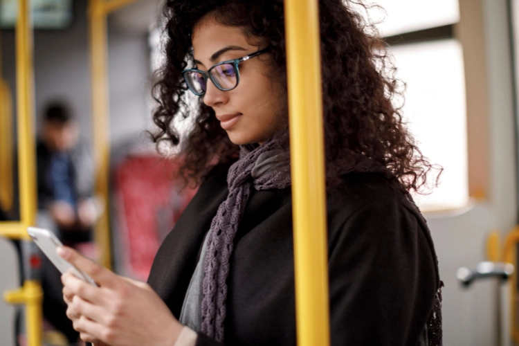 A young woman with dark, curly hair and blue glasses looks intently at her smartphone while riding public transportation. She is wearing a dark coat and a gray scarf.