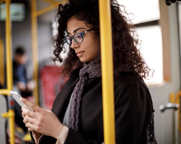 A young woman with dark, curly hair and blue glasses looks intently at her smartphone while riding public transportation. She is wearing a dark coat and a gray scarf.