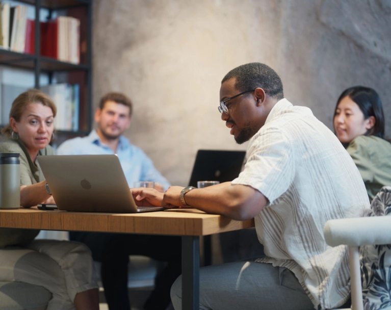 A diverse group of four people sits around a table in a modern office, working on laptops. The man in the foreground is wearing glasses and a light-colored shirt, focusing on his computer.