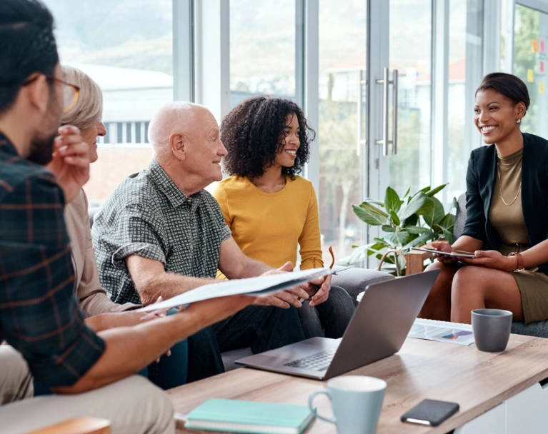 A diverse, intergenerational group sits in a brightly lit room having a discussion around a coffee table with a laptop. A Black woman on the right is smiling at the group while holding a tablet.