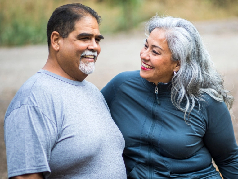 A senior Hispanic couple, both smiling, stand outdoors. The man is wearing a gray t-shirt and has a gray mustache, and the woman has long gray hair and a blue athletic jacket.