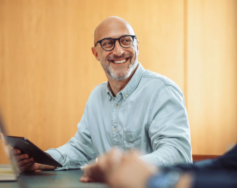 A smiling, bald man with glasses and a beard sits at a conference table, holding a tablet and looking towards his colleagues in a professional setting.