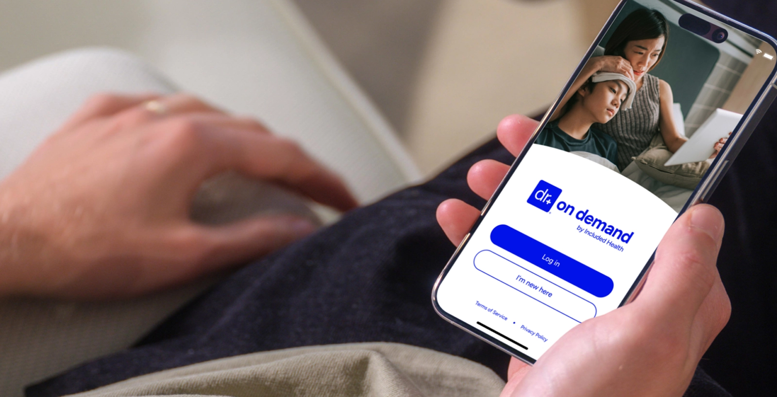 A close-up of a hand holding a smartphone displaying the login screen for the "Dr On Demand by Included Health" mobile app. The background of the screen shows a mother applying a compress to her child's forehead during what appears to be a virtual visit.