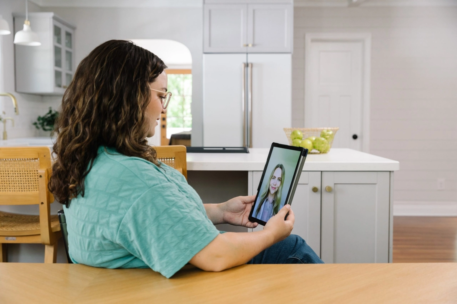 A woman with curly hair and glasses sits in her kitchen, holding a tablet for a virtual care video appointment with a doctor.