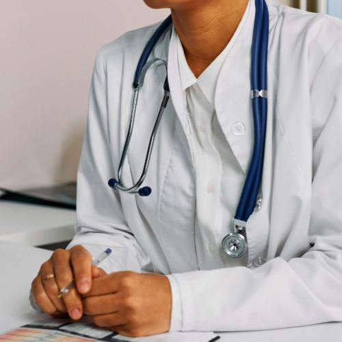Close-up shot of a medical professional in a white lab coat with a blue stethoscope around their neck. Their hands, holding a pen, rest on a desk.