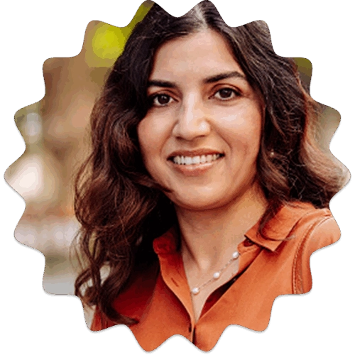 A smiling woman with dark, wavy hair and an orange shirt stands outdoors, looking directly at the camera.