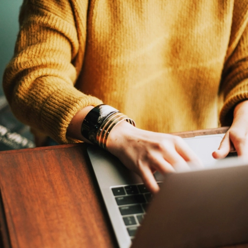 Close-up view of a person wearing a yellow knitted sweater and gold bracelets, typing on a silver laptop keyboard placed on a dark wood table.