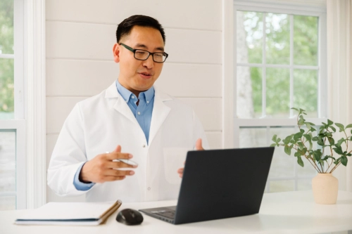A medical professional, an Asian man wearing a white lab coat and glasses over a blue shirt, sits at a desk in a bright room, gesturing while conducting a virtual consultation on a laptop.