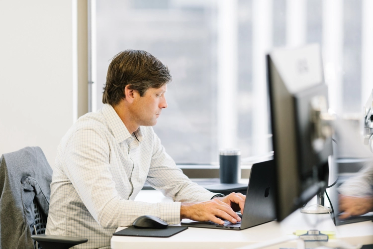 A focused man in a checkered shirt sits at an office desk near a window, typing on a black laptop with two monitors visible.
