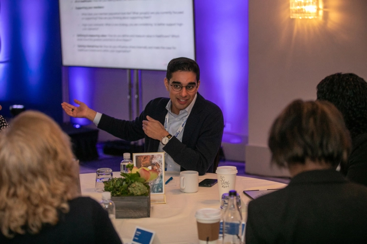 A man wearing glasses and a suit jacket gestures while speaking to a diverse group of colleagues seated around a round conference table during a morning business session.