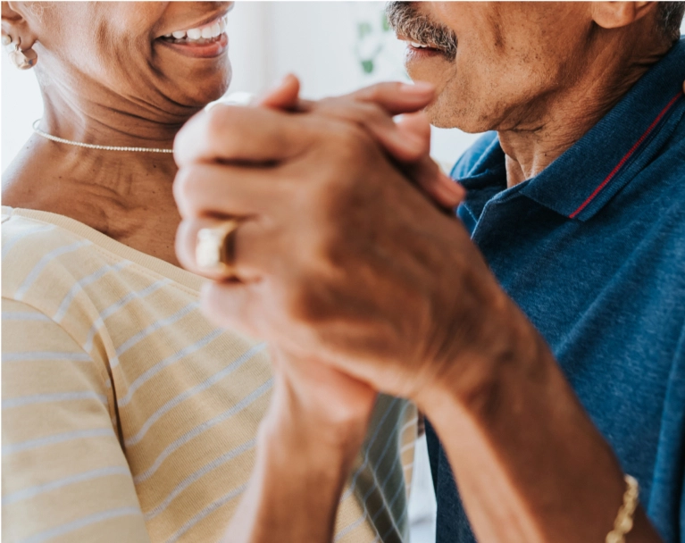 A close-up of a joyful senior Black couple dancing and holding hands. The man has a mustache and the woman is smiling, wearing a beaded necklace.