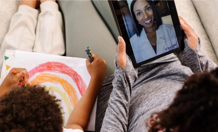 A mother sits with her child, who is coloring a rainbow. The mother is holding a tablet that displays a smiling female doctor on a video call for a virtual visit.