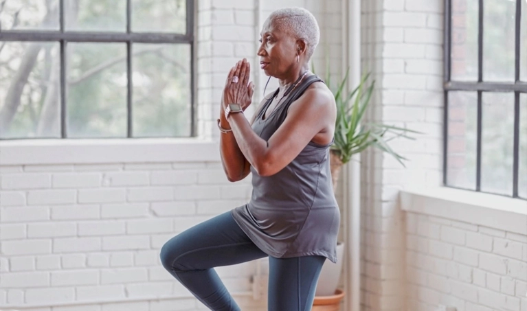 A senior woman with short gray hair performs a tree yoga pose, standing on one leg with her hands pressed together. She is wearing exercise clothes and is near large industrial windows.