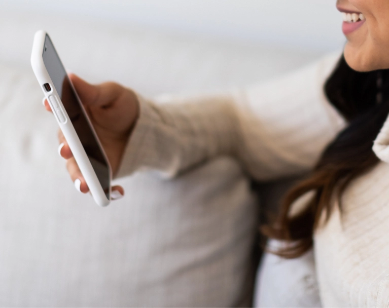 Close-up of a woman's hand with white nail polish holding a white smartphone in a well-lit, cozy indoor setting.