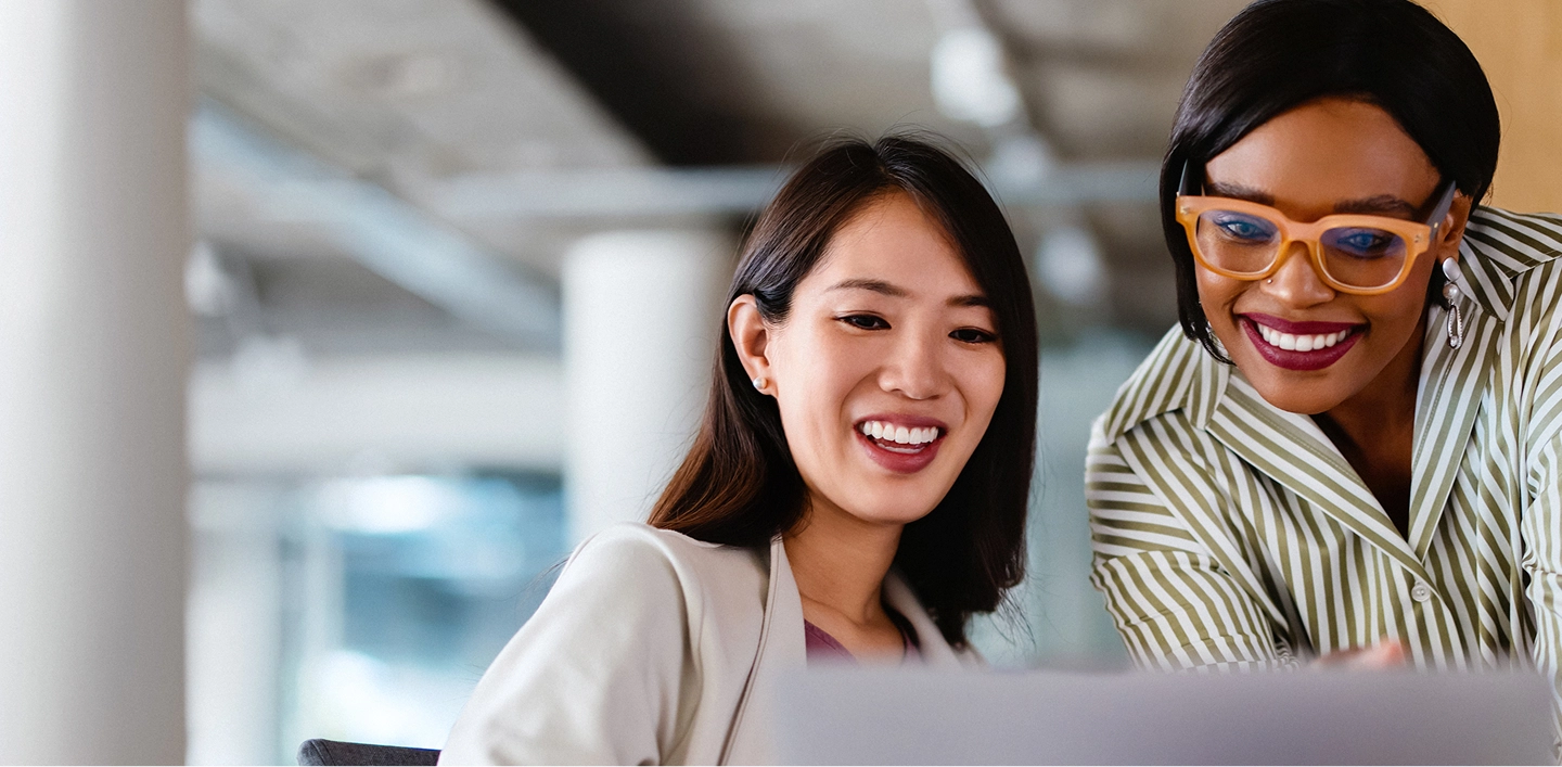A close-up of two diverse professional women in an office setting, both smiling and looking down at a laptop screen. The woman on the right is wearing large orange glasses and a striped shirt.