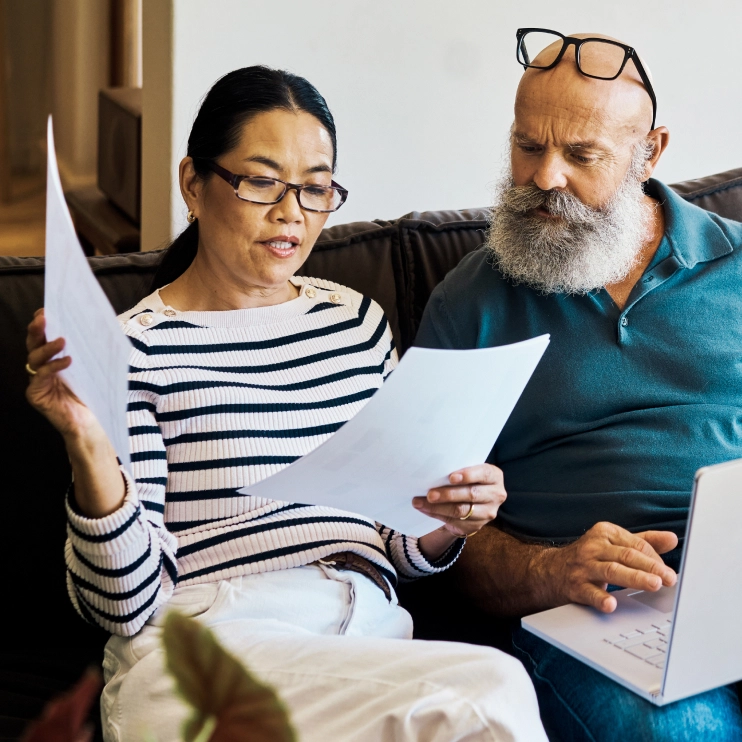 A couple reviewing paperwork at home on a couch. The woman is holding documents and wearing glasses, and the man with a long white beard has a laptop on his lap.
