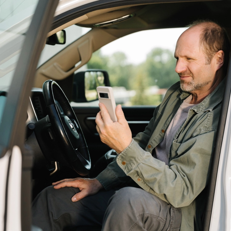 A man sitting in the driver's seat of a truck, looking at his smartphone. Overlaid on the image is an app message bubble that reads, "Ready to check-in? This will only take a minute."