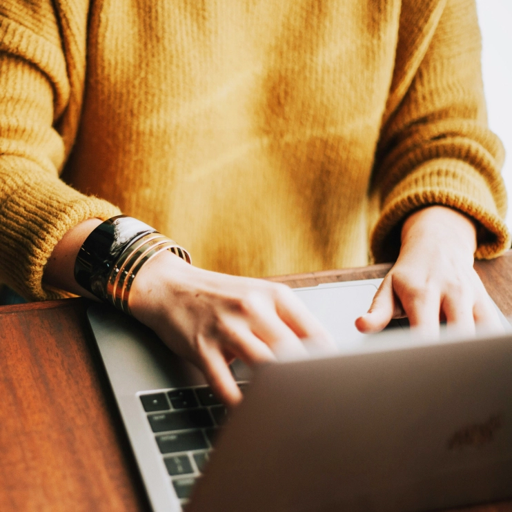 A close-up of a person wearing a mustard yellow sweater and black and gold bracelets, typing quickly on a laptop keyboard.