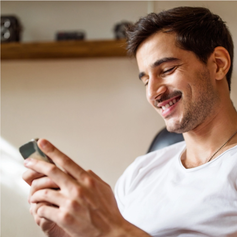 A young man with a mustache smiles while looking down and interacting with his smartphone.