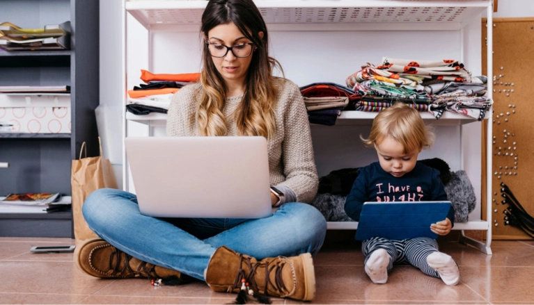 A young woman wearing glasses and a sweater sits cross-legged on the floor, working on a laptop, next to a small child who is distracted by a tablet. Shelves with various items are in the background, symbolizing remote work and family life.