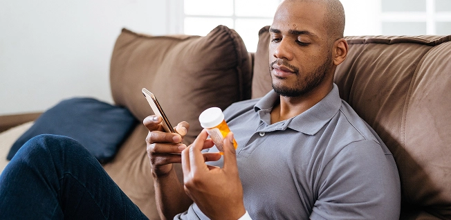 A man with a shaved head sits on a couch, looking at a prescription medicine bottle in one hand and his smartphone in the other, likely checking dosage or cost.