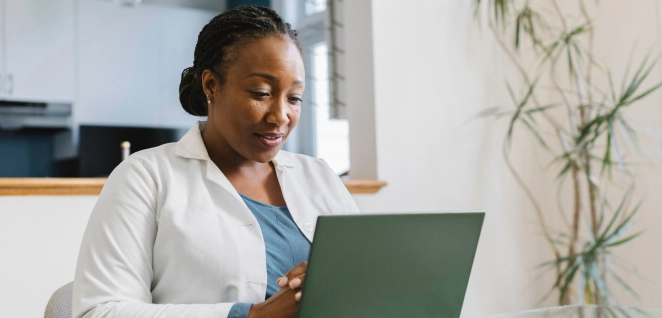 A Black woman in a white lab coat conducts a virtual patient consultation from her home office, looking intently at her open laptop.
