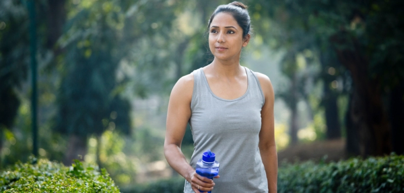 A woman in athletic wear walks outdoors in a park, holding a blue water bottle, symbolizing exercise and healthy living.