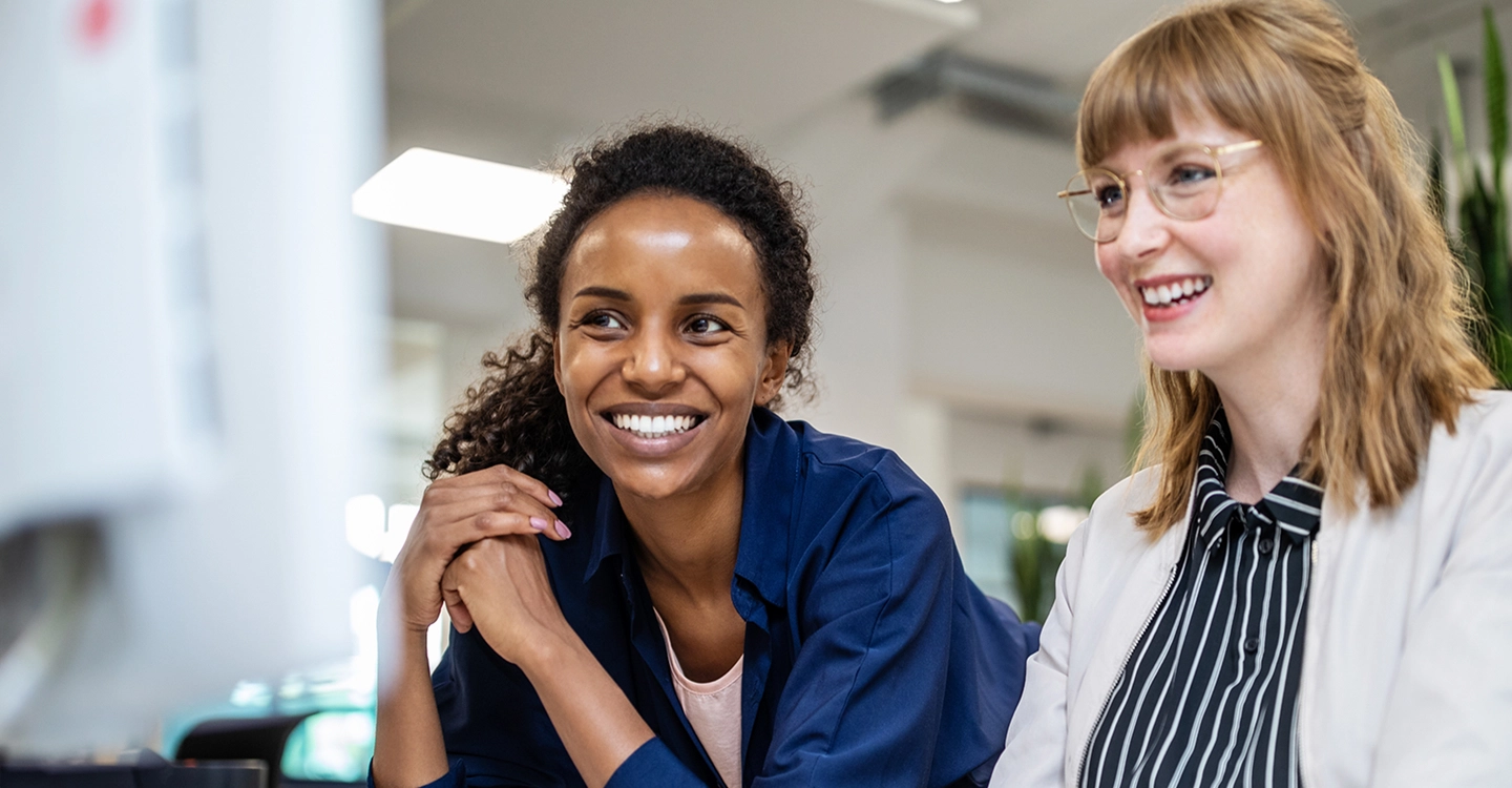 Two smiling professional women, one Black and one White, looking together at a computer screen in an office setting.