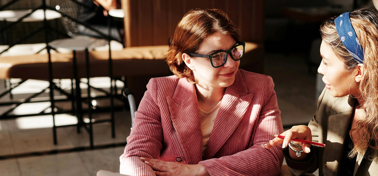 Two women are having a discussion in a bright, modern cafe. One woman, wearing a pink blazer, listens as the other woman, wearing a blue bandana, speaks and gestures with a pen.