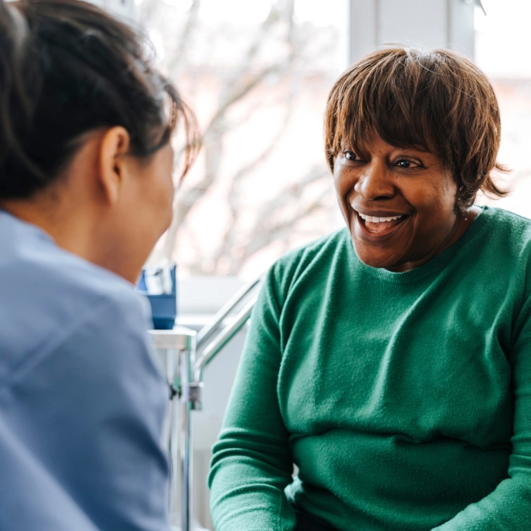 A Black woman wearing a green sweater laughs enthusiastically while speaking to a healthcare provider in a blue uniform, likely during a successful consultation.