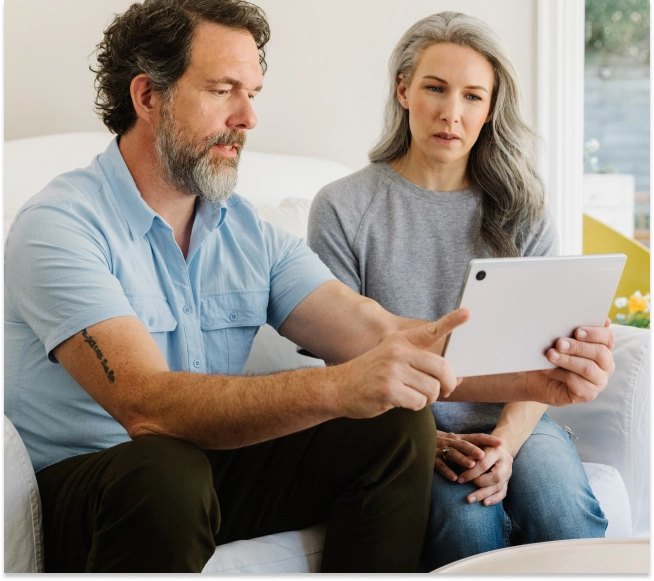 A couple sits together on a sofa, looking down at a tablet computer held by the woman, possibly engaging in a joint virtual care appointment.