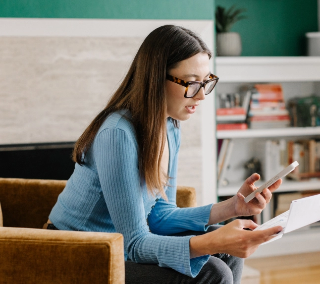 A woman with glasses and long brown hair sits in an armchair, looking intently at a document (likely a medical bill) in one hand while holding her smartphone in the other, symbolizing complex billing navigation.