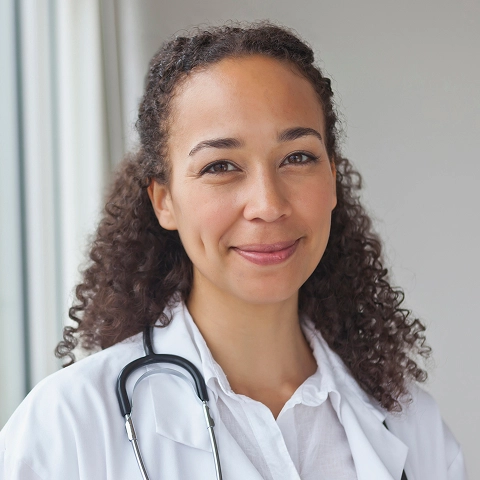 A portrait of a smiling female doctor with curly brown hair, wearing a white lab coat and stethoscope, standing next to a window.