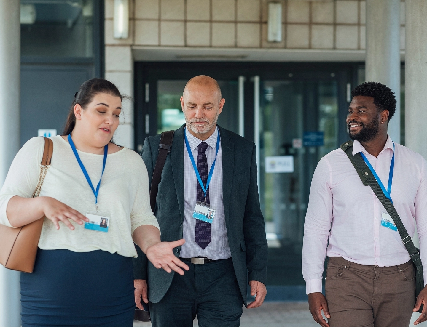 Three professionals wearing lanyards and ID badges stand together outside a building entrance. A woman on the left gestures as she speaks, while a bald man and a Black man listen and smile.