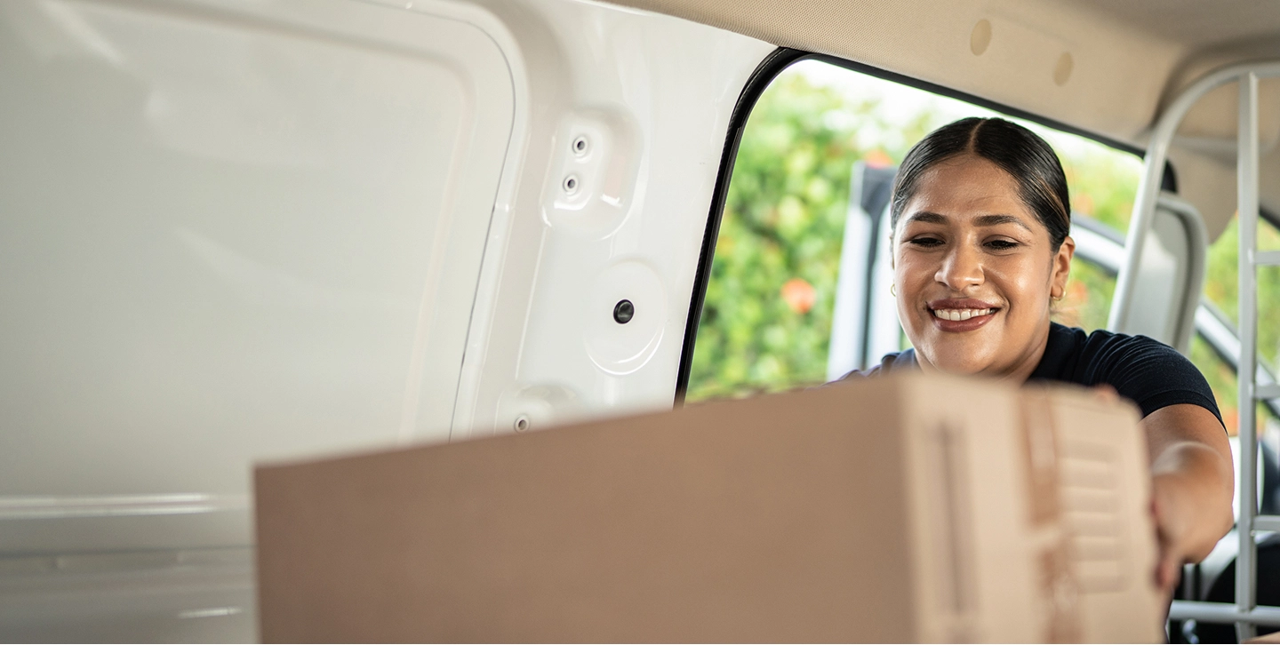 A smiling woman with dark hair pulled back, wearing a dark t-shirt, loads a large cardboard box into the open back of a white delivery van.